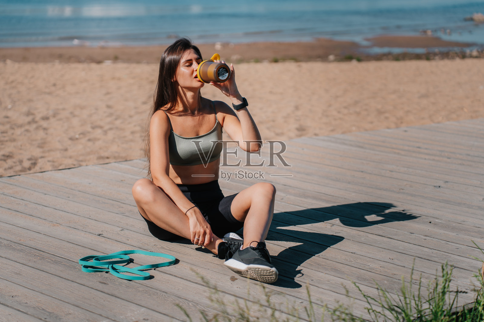 一个放松的女人在锻炼后喝着水，坐在海滩的木板路上，旁边有阻力带照片摄影图片
