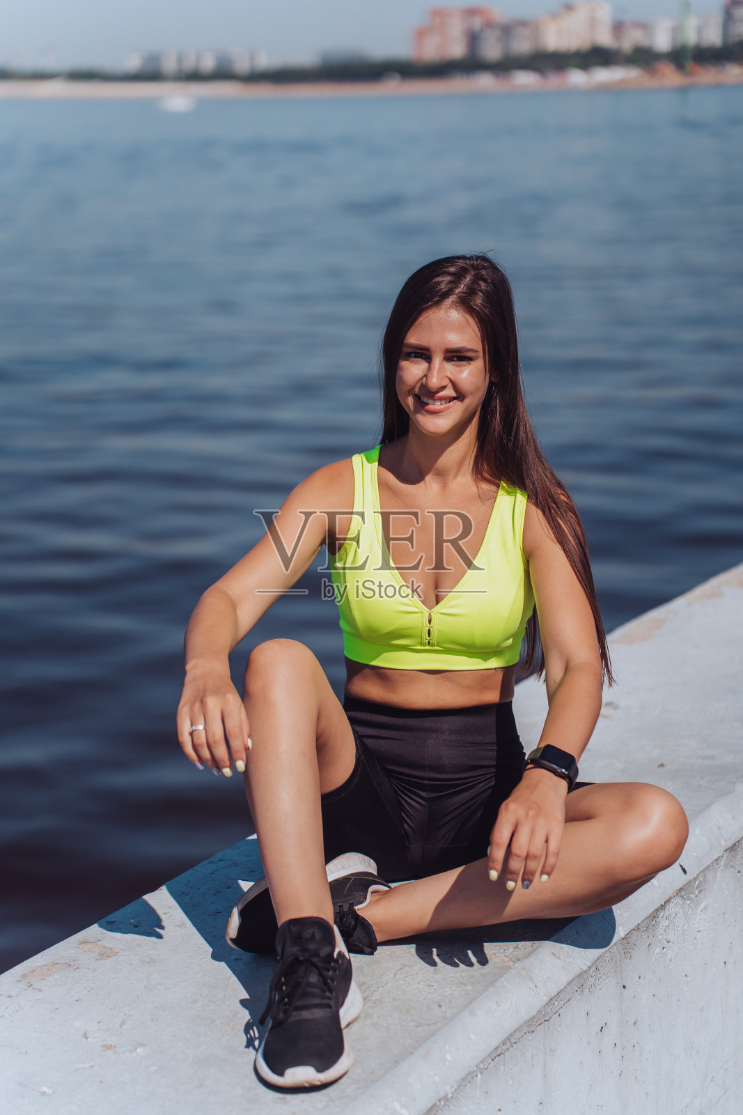 穿着鲜艳运动服的微笑女子坐在水边，享受阳光，手腕上戴着健身追踪器。照片摄影图片