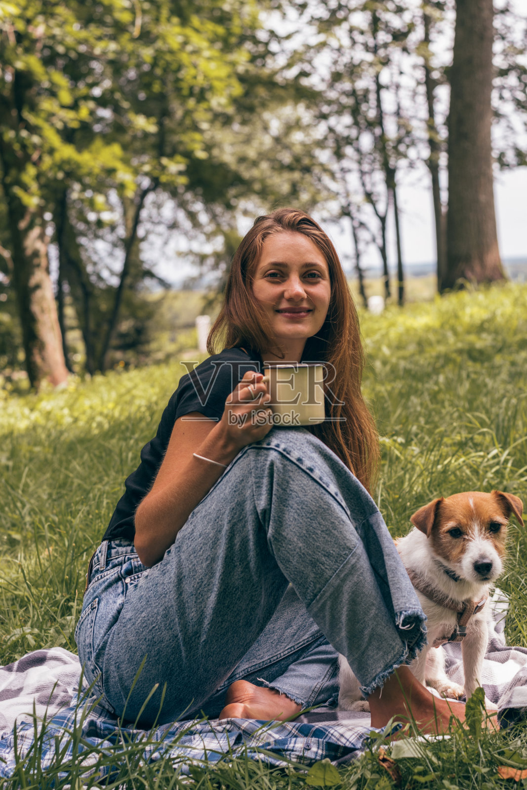 一位乐观的年轻女子牵着她的狗在公园里散步。一个女人用保温瓶喝茶。一只杰克罗素梗狗坐在她旁边照片摄影图片