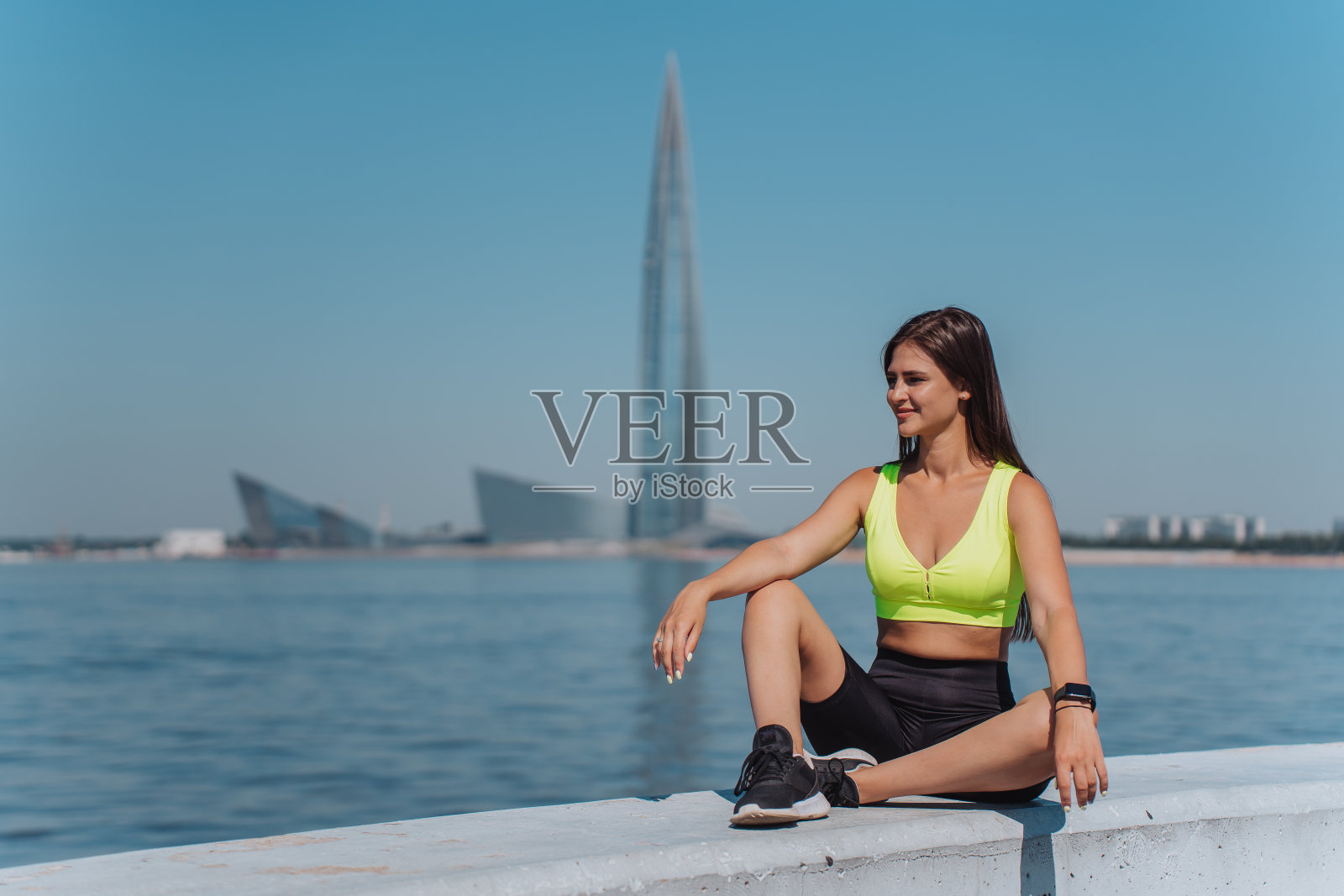 运动的黑发欧洲女孩慢跑后放松坐在堤岸闭着眼睛穿着运动服在阳光明媚的日子。健康的女人在现代摩天大楼的映衬下享受阳光。健身、锻炼。照片摄影图片