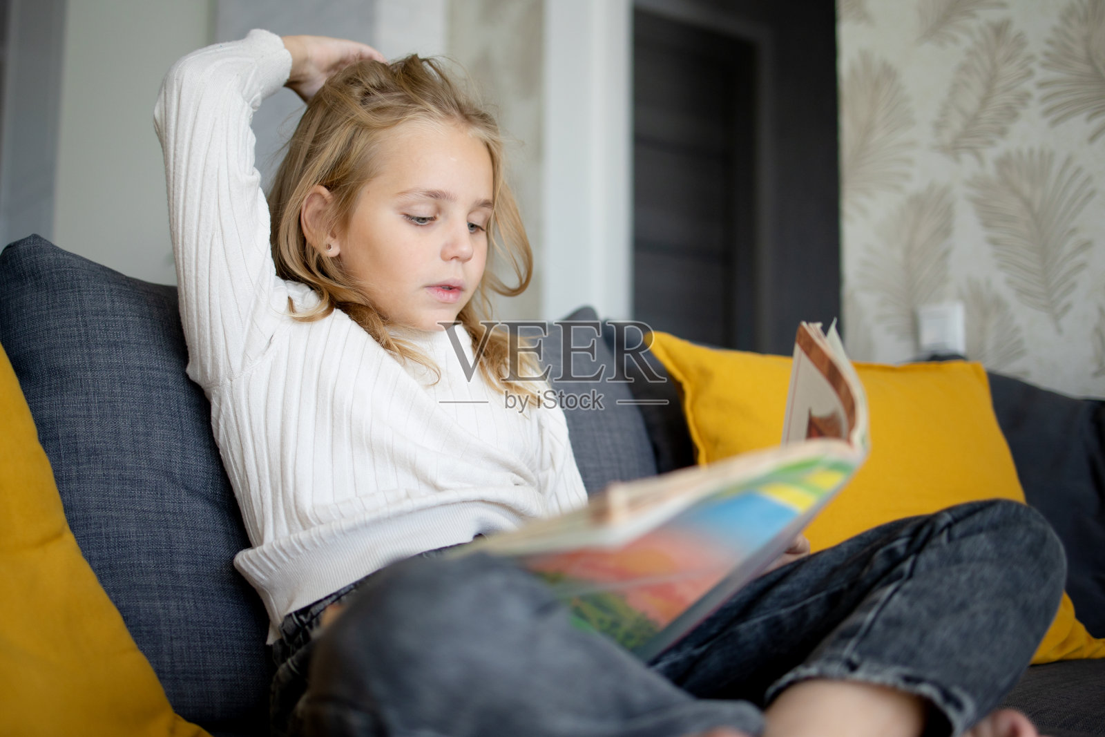 一个金发小女孩坐在家里的沙发上，沉浸在一本有趣的书的迷人情节中。照片摄影图片