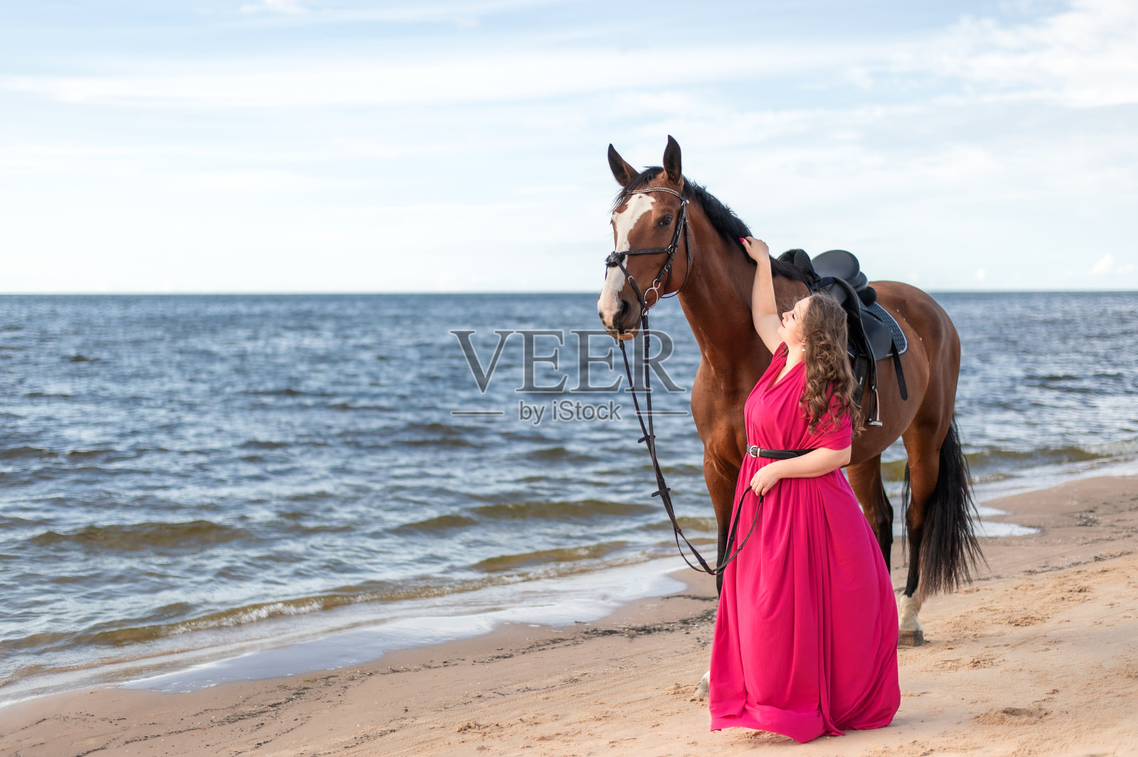 穿着粉色连衣裙的迷人年轻女子和她的马在沙滩上照片摄影图片