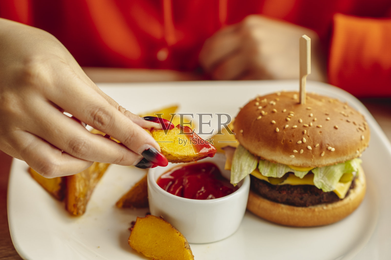 白盘子上有汉堡、土豆片和酱汁。不认识的女人拿起一块土豆，蘸上酱汁，坐在餐馆里吃。食品的概念。关闭了。照片摄影图片