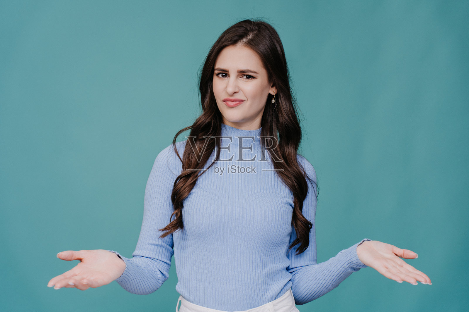 不开心的黑发年轻女子穿着淡紫色毛衣，困惑地摊开双手，站在蓝绿色演播室背景前，不安。人们真诚的情感。样机照片摄影图片