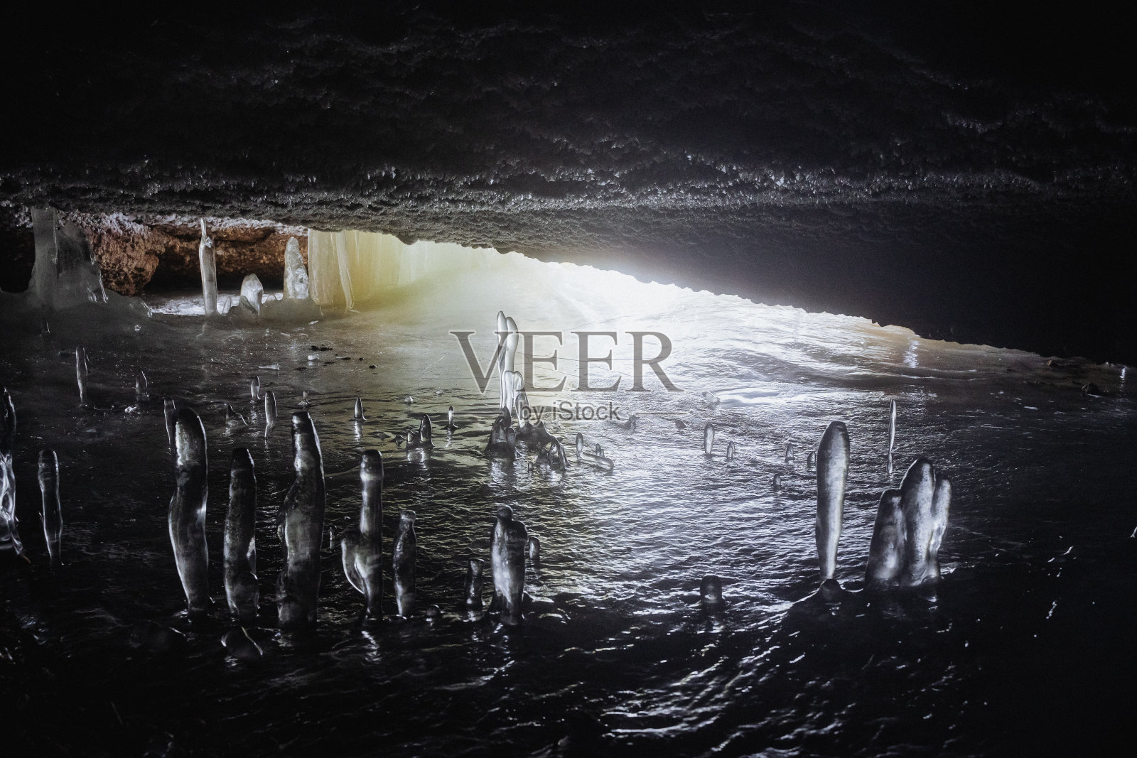 地下有一个覆盖着冰的洞穴，冰石笋生长在洞穴里，地下有冰冻的水，尽头有光出口。照片摄影图片