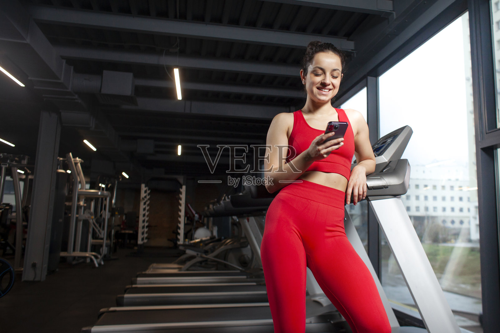 穿着红色运动服的运动女孩在健身房使用智能手机，微笑着，迷人的苗条女人在训练中拿着手机照片摄影图片