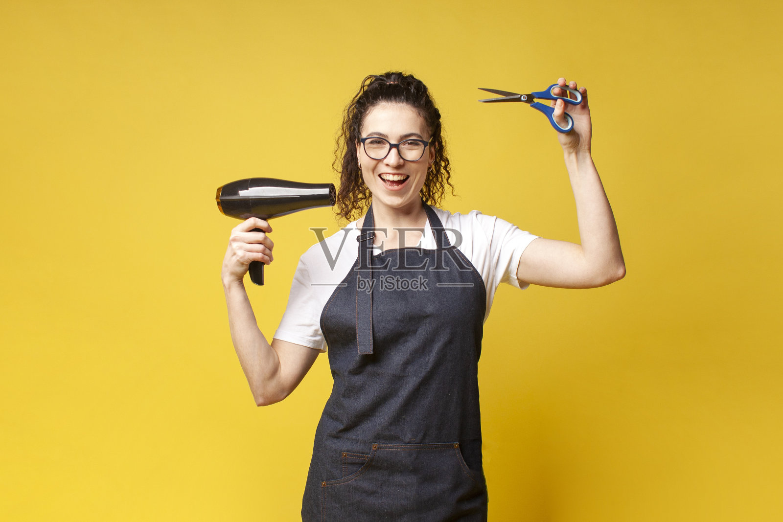 穿着制服的年轻美发师拿着吹风机和剪刀，在黄色背景上跳舞照片摄影图片
