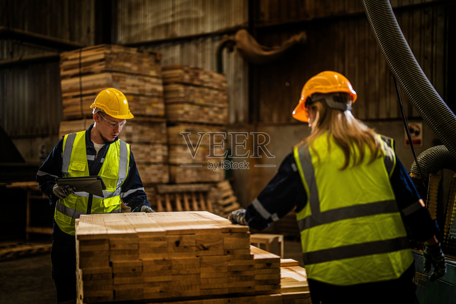 班组木工穿着安全制服，戴安全帽，在生产车间检查木制品的质量。男女工人在黑暗的仓库工业木材。照片摄影图片