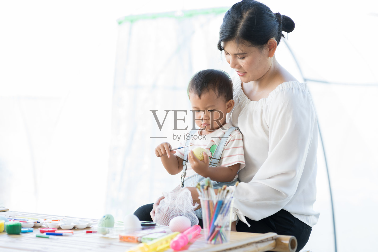 亚洲母亲和幼小的儿子在课堂上用毛笔画鸡蛋。快乐的亚洲妈妈抱着他的小儿子，教他用水彩画鸡蛋照片摄影图片