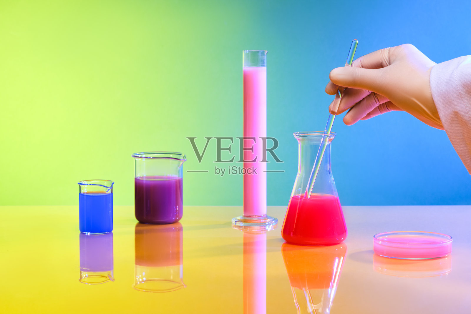 一只手拿着一根玻璃棒，在一个装有红色液体的埃伦迈尔烧瓶里搅拌，旁边是一个装有彩色液体的玻璃器皿，背景颜色从黄色变为蓝色。照片摄影图片