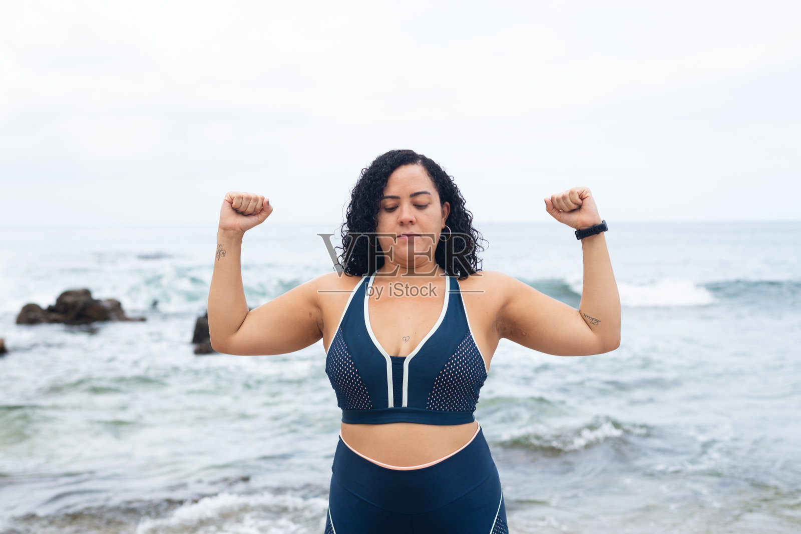 健身的女人，站着，用她的手臂在沙滩上做运动，对着大海和多云的天空。照片摄影图片