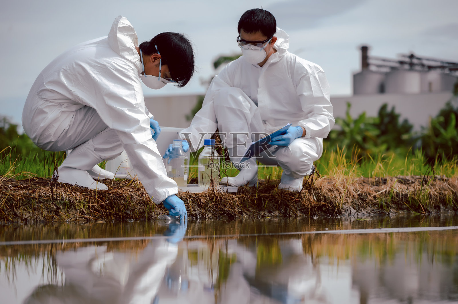 工厂的科学家或生物学家在农田附近的天然水源采集水样时穿着防护服。化学防护的概念可能会渗入天然水源。照片摄影图片