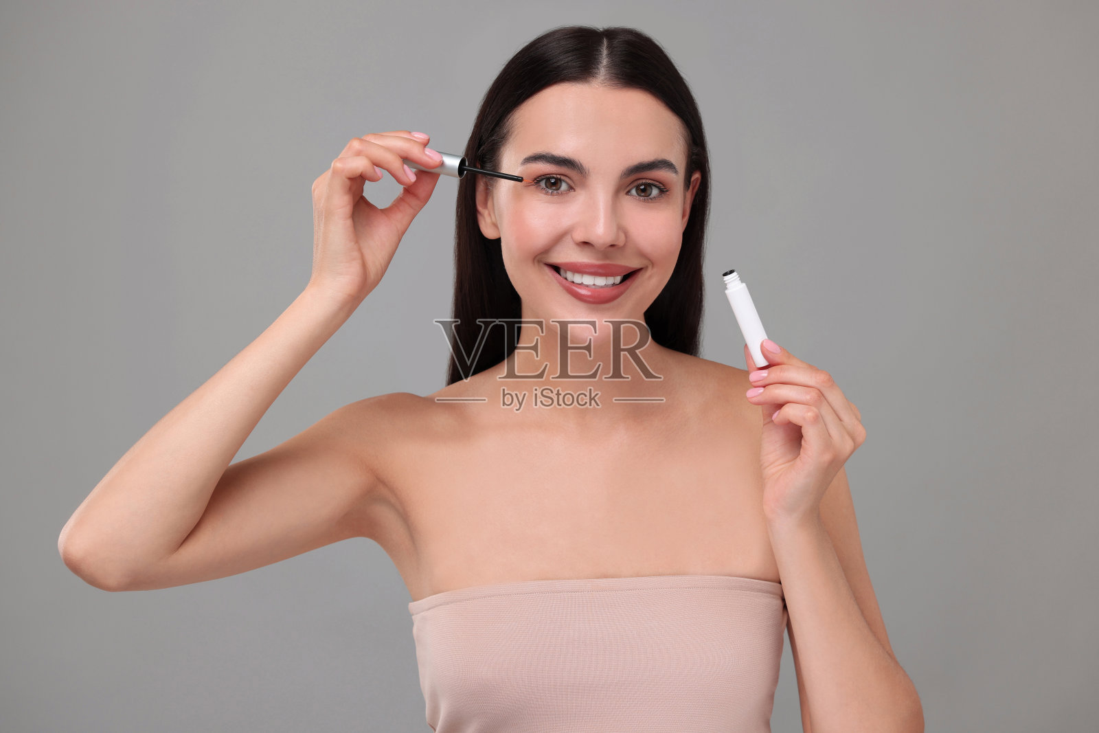 美丽的女人把精华液涂在灰色背景的睫毛上。美容产品照片摄影图片