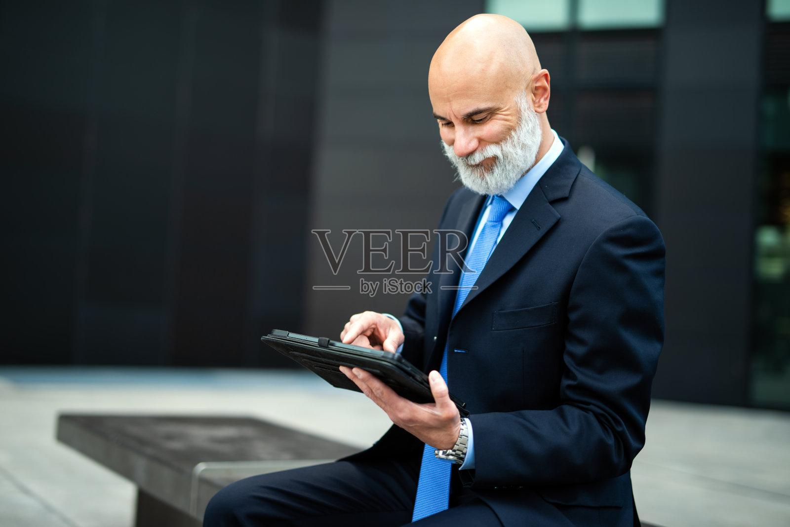 时髦的经理坐在现代城市户外的长凳上使用他的平板电脑照片摄影图片