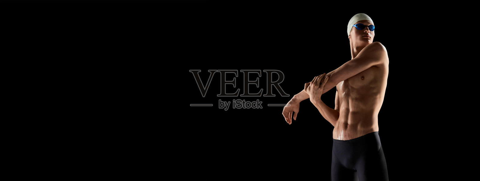 肌肉发达，体格健壮的年轻人，游泳者，戴着泳镜和泳帽，在黑色背景下游泳前伸展肌肉。照片摄影图片