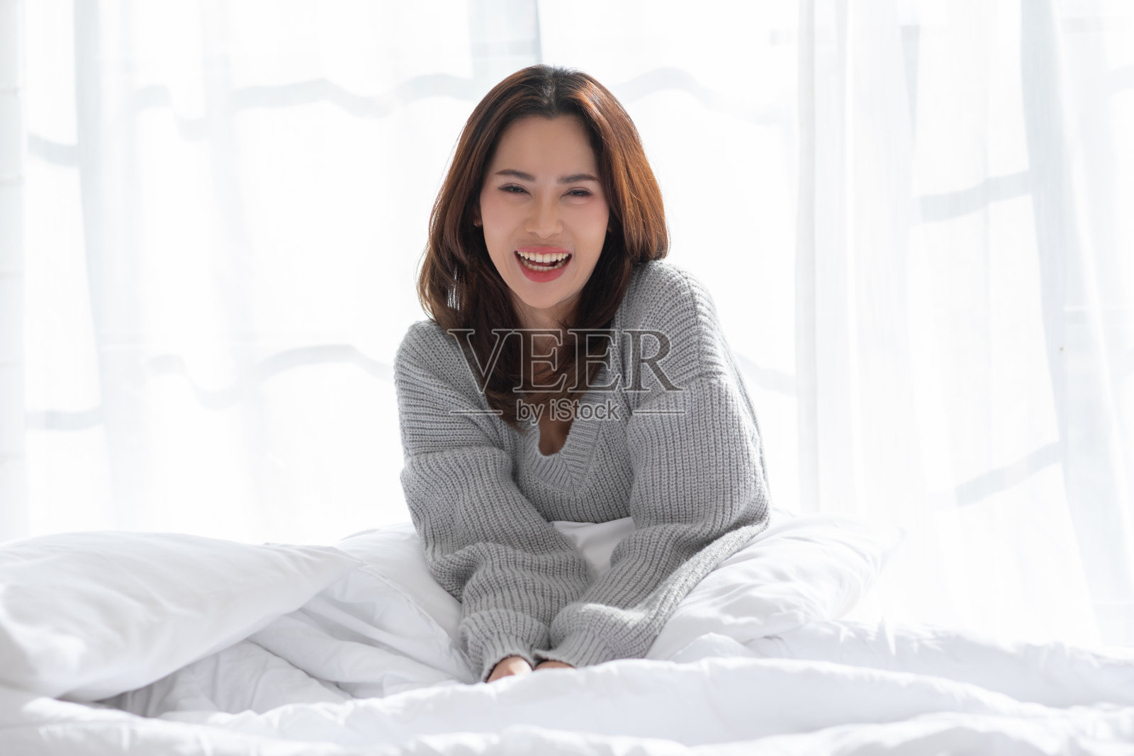 迷人的亚洲年轻女子坐在床上看着相机，微笑着幸福和舒适的家。可爱的女孩微笑，交叉手臂自信，放松在卧室里照片摄影图片