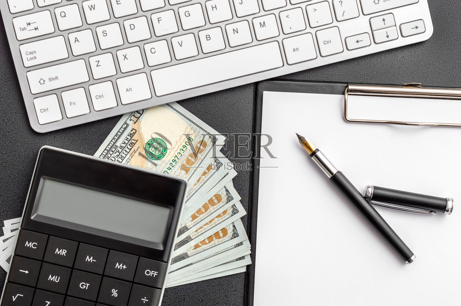 黑色桌子上有带钱的计算器、剪贴板和电脑键盘。照片摄影图片