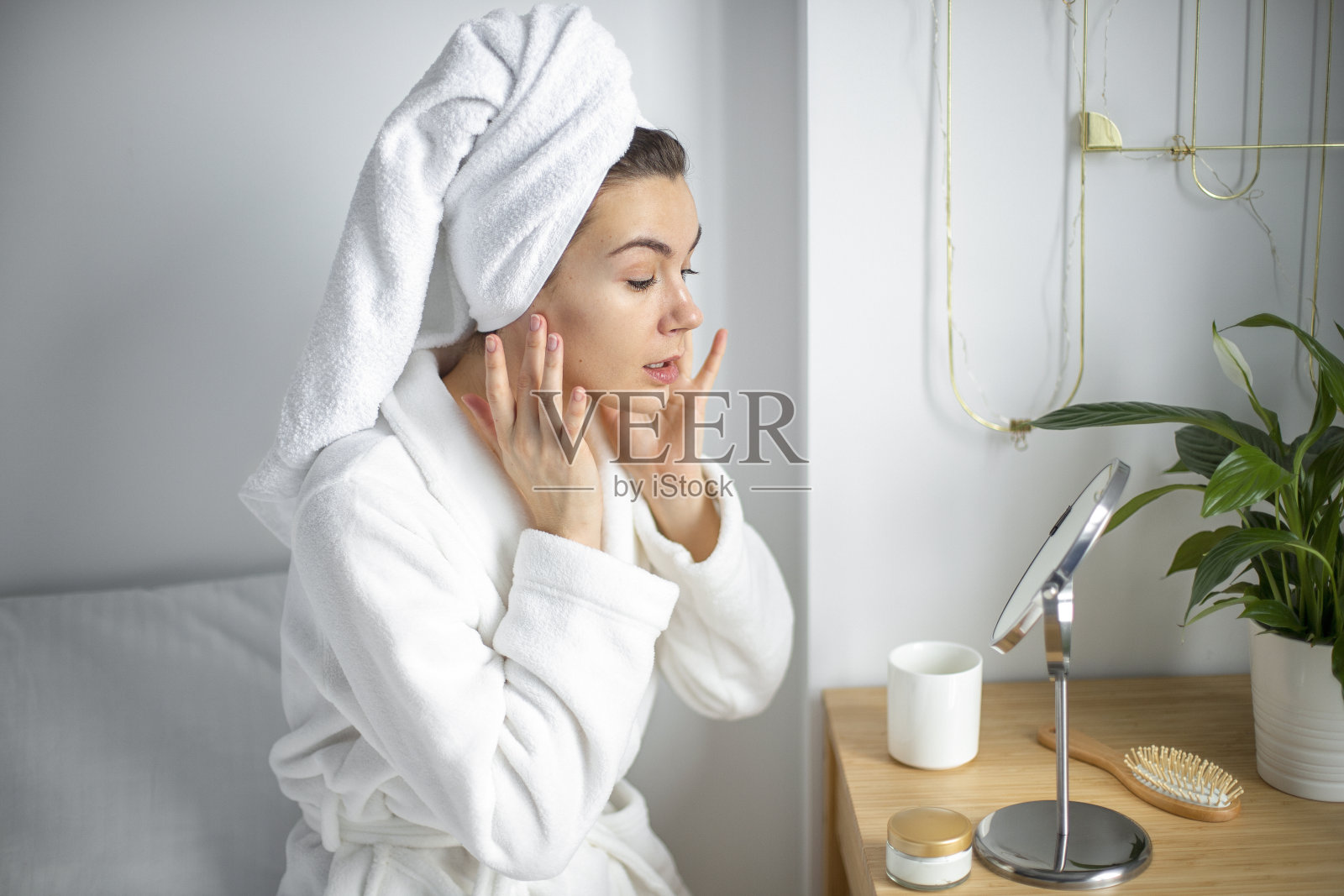 穿着白色浴袍，头裹毛巾的美女，在脸上抹上面霜，温柔地按摩脸部，享受愉悦，享受清晨时光，自我护理理念照片摄影图片