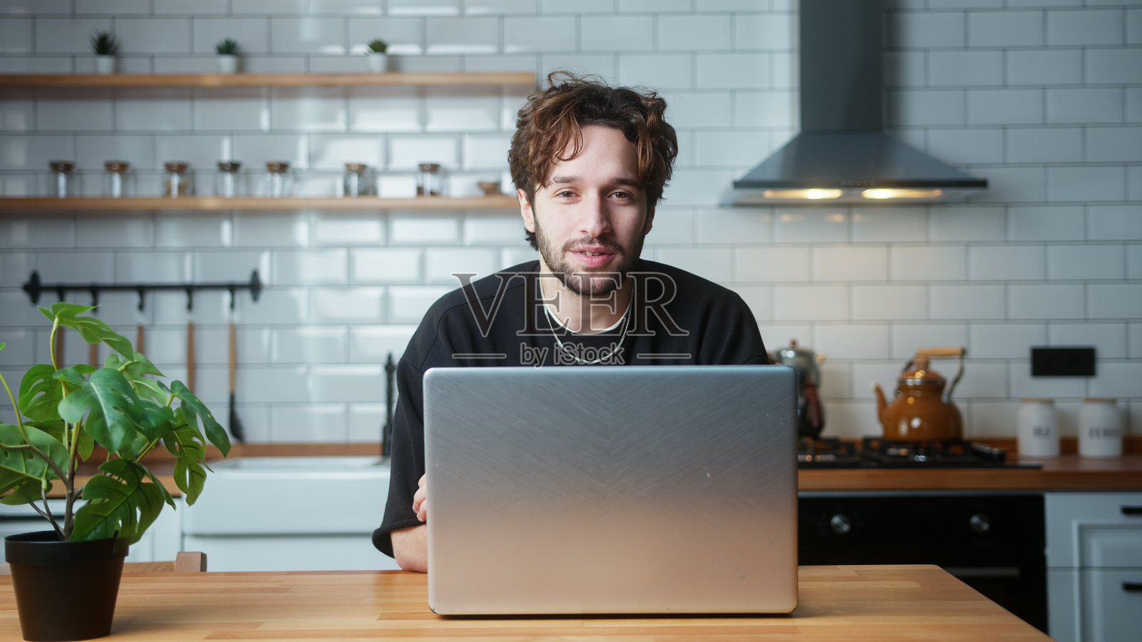 年轻卷发男子坐在家里的现代厨房里看着相机的肖像照片摄影图片