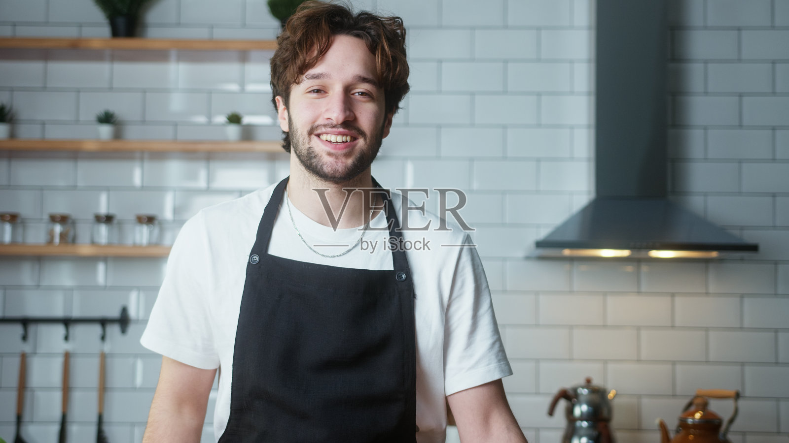 年轻微笑男子的肖像，卷发，系着围裙，看着相机在家里的厨房照片摄影图片
