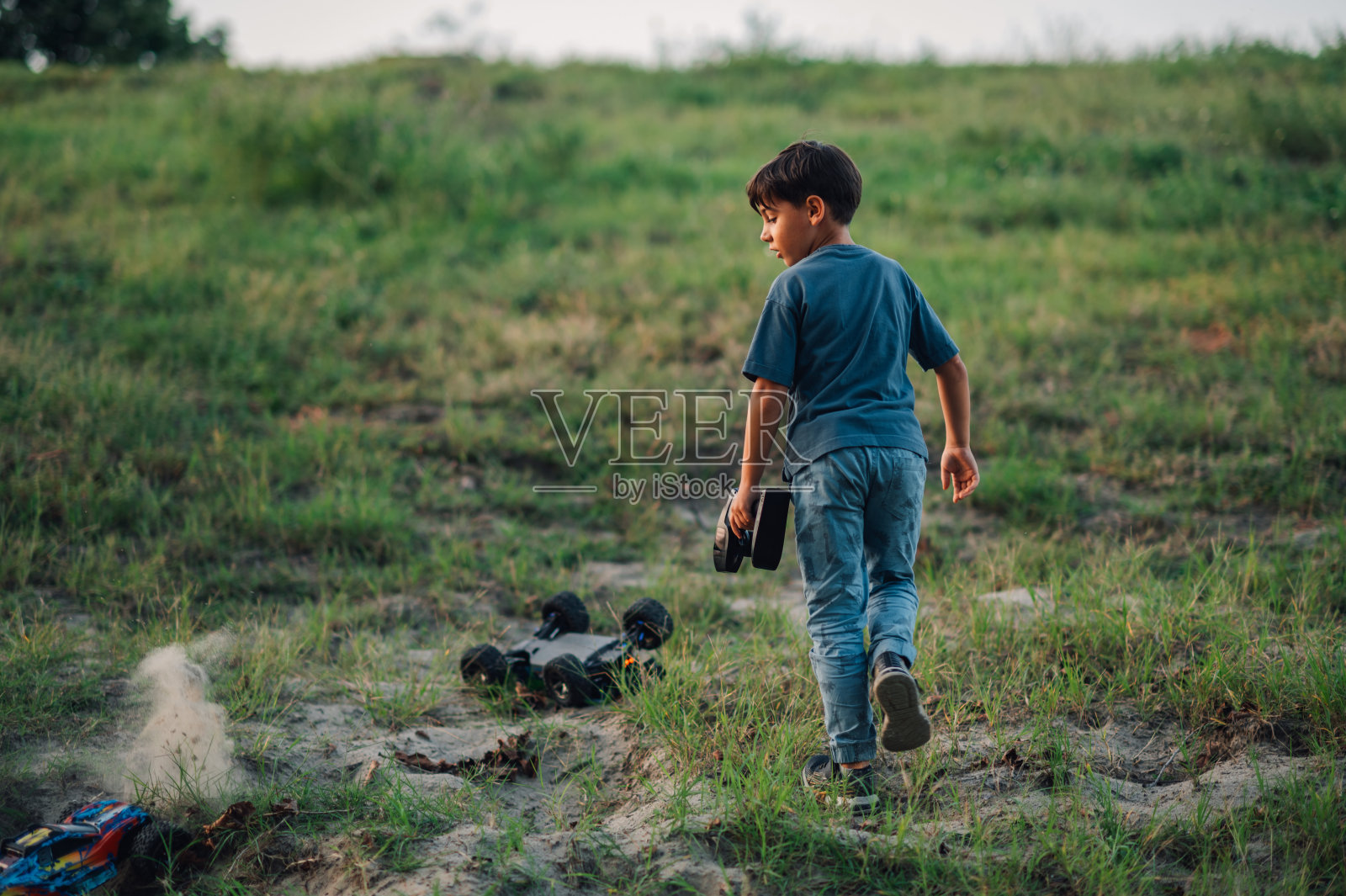 一个小男孩跑向他打翻的玩具车，在大自然的草地上。照片摄影图片