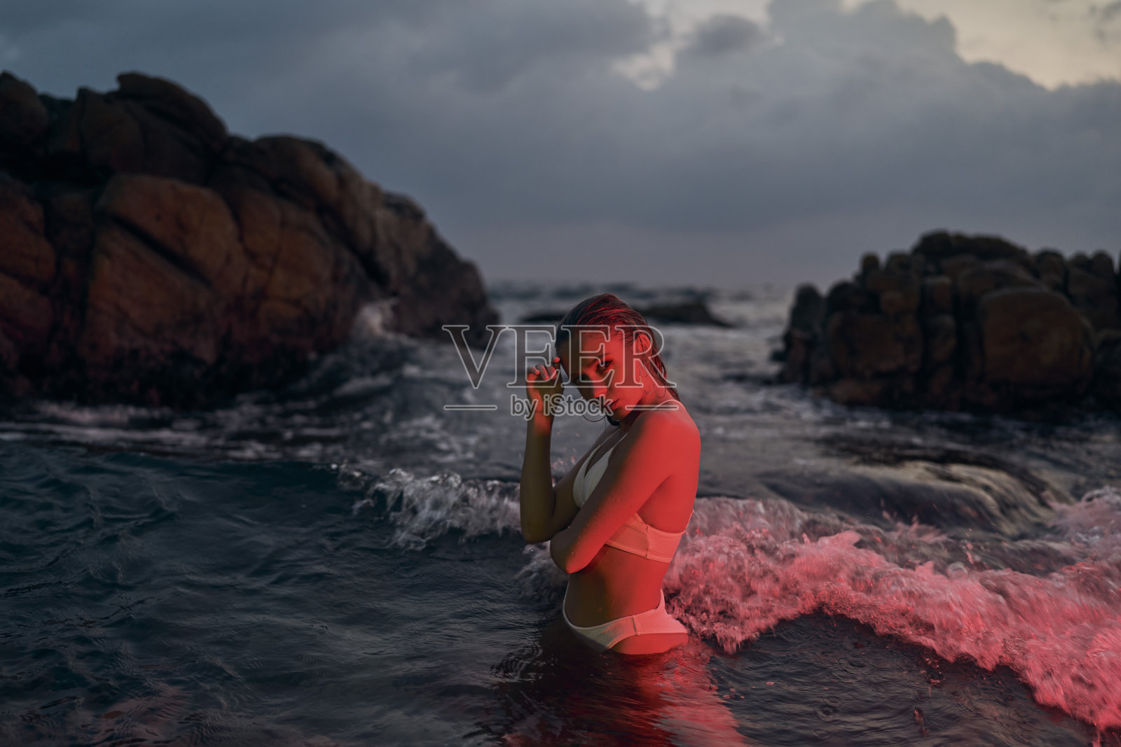 迷人的女人喜欢晚上在红色灯光照耀下的海洋里游泳。穿着泳装的时尚女性体验夜间水上冒险。优雅的淑女在发光的大海，美丽的理念在大自然。照片摄影图片