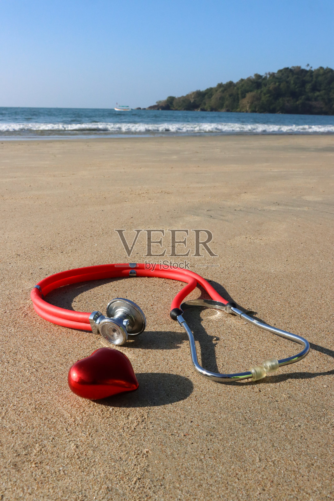 模特形象，听诊器在阳光明媚的沙滩上的塑料红心，退潮时的海浪，大海和海岸线，无压力的健康和幸福概念，重点放在前景上照片摄影图片