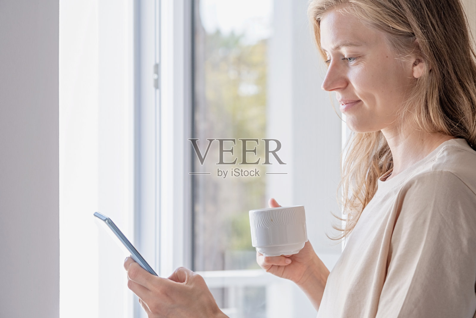 女子站在窗边用手机端着咖啡杯照片摄影图片