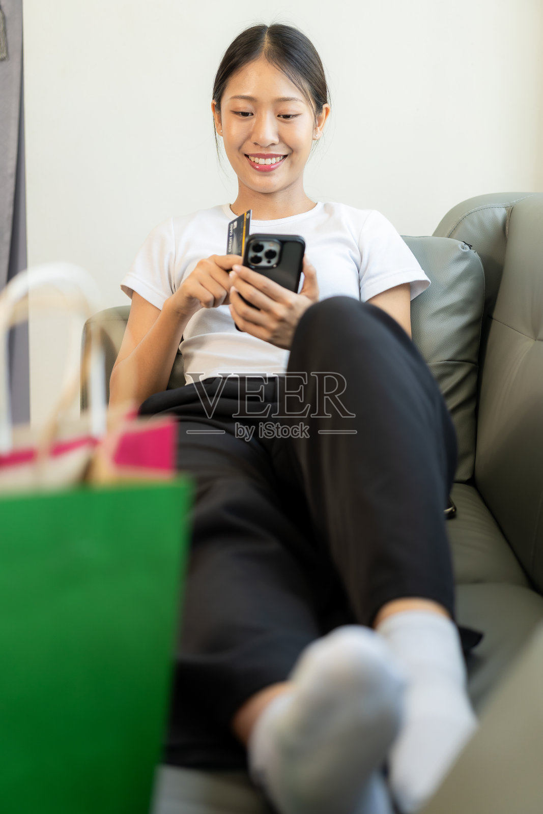 开朗的亚洲女性使用智能手机和笔记本电脑，网上购物，持有信用卡，支付，坐在家里的沙发上，通过网上银行应用程序支付概念。照片摄影图片