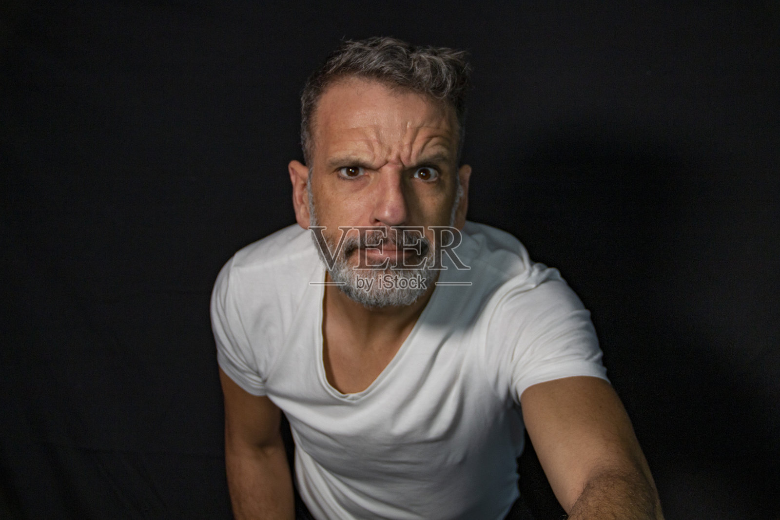 一个成熟男人的肖像，留着灰胡子，戴着牙套，用手打手势，穿着黑色背景上的白色t恤。照片摄影图片