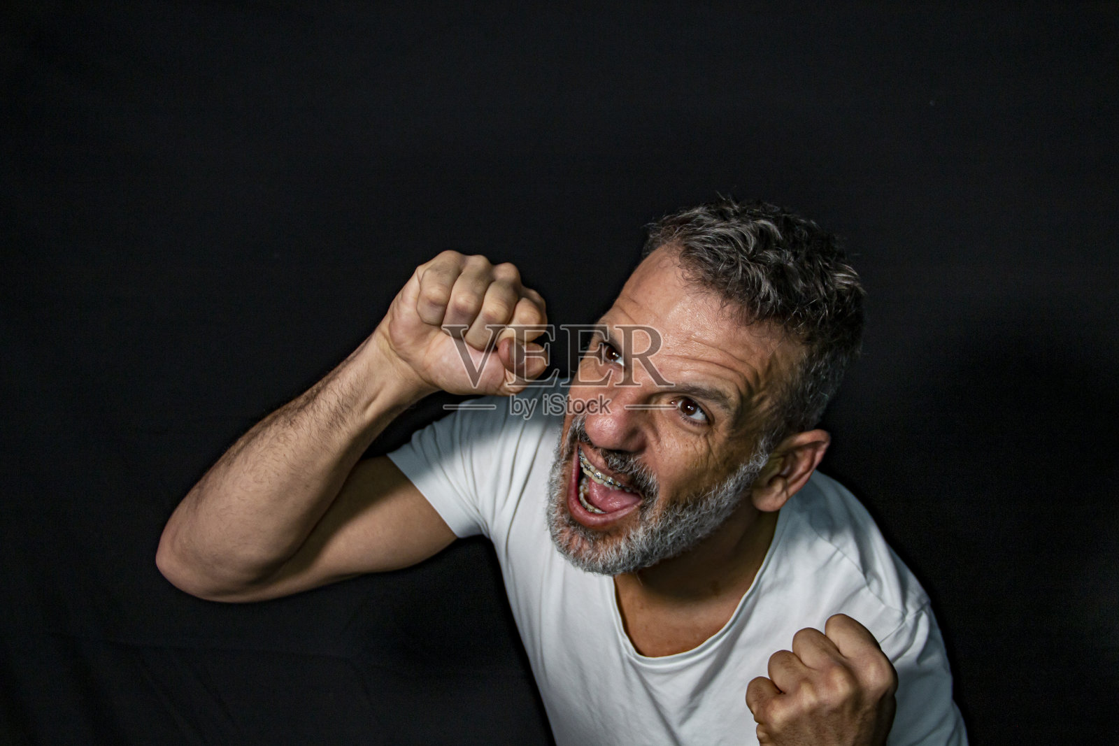 一个成熟男人的肖像，留着灰胡子，戴着牙套，用手打手势，穿着黑色背景上的白色t恤。照片摄影图片