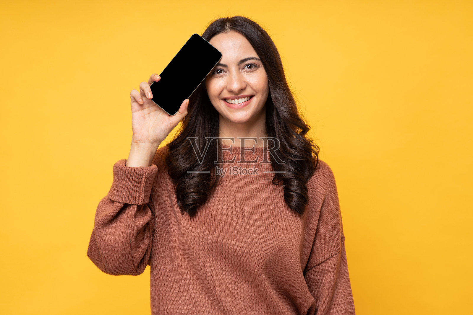 照片中，美丽快乐的年轻女子穿着运动衫，孤立地站在黄色背景上，展示了手机的空白屏幕照片摄影图片