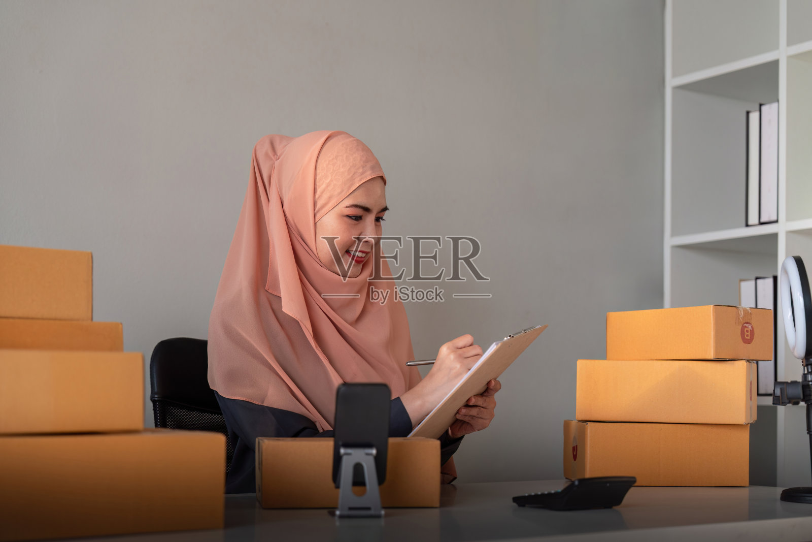 穆斯林妇女在家拿着盒子在网上卖东西。在线销售，接受客户的订单。中小企业经营理念。包裹递送。穆斯林妇女在家使用智能手机和笔记本电脑照片摄影图片