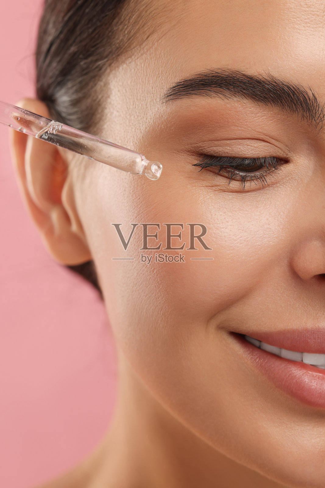 美丽的年轻女子将精华液涂抹在她的脸上，粉红色的背景，特写照片摄影图片