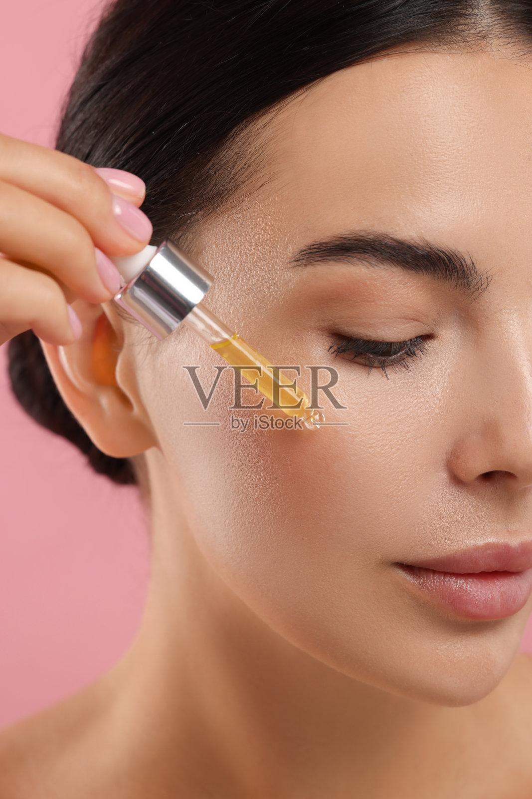 美丽的年轻女子将精华液涂抹在她的脸上，粉红色的背景，特写照片摄影图片