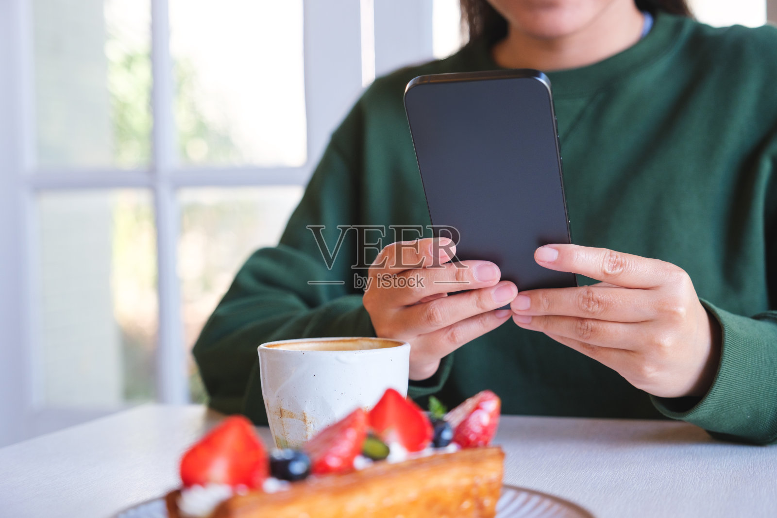 一名年轻女子手持并使用手机的特写照片，桌上放着咖啡杯和蛋糕照片摄影图片