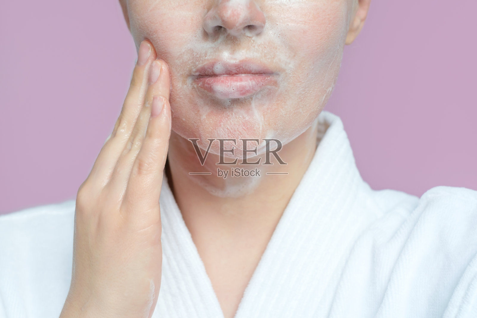 年轻女子正在用洁面泡沫擦拭脸部特写，背景为丁香色。照片摄影图片