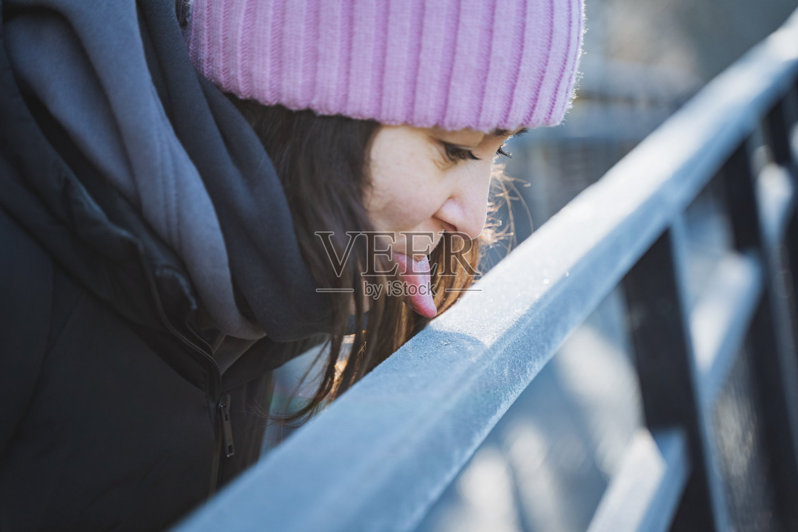 一个女孩用她的舌头舔着冰冻的栏杆，一个年轻女子在新鲜空气中游荡，用她的舌头触摸着冰冷的表面。照片摄影图片