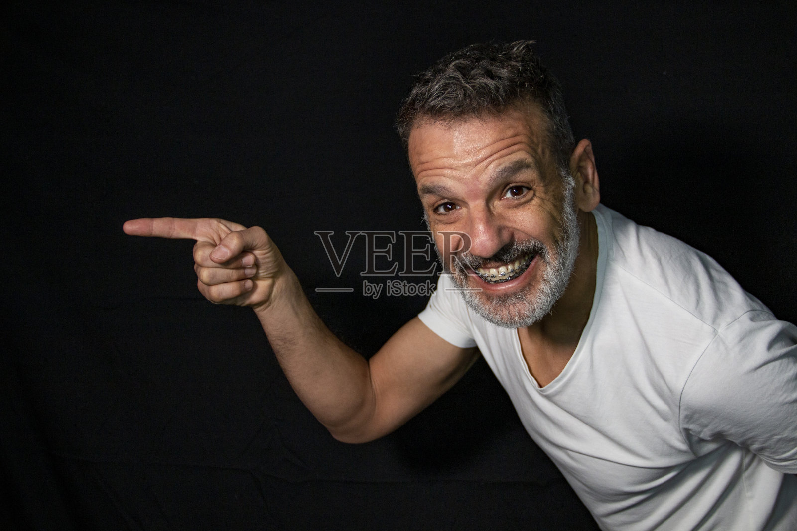 一个成熟男人的肖像，留着灰胡子，戴着牙套，用手打手势，穿着黑色背景上的白色t恤。照片摄影图片
