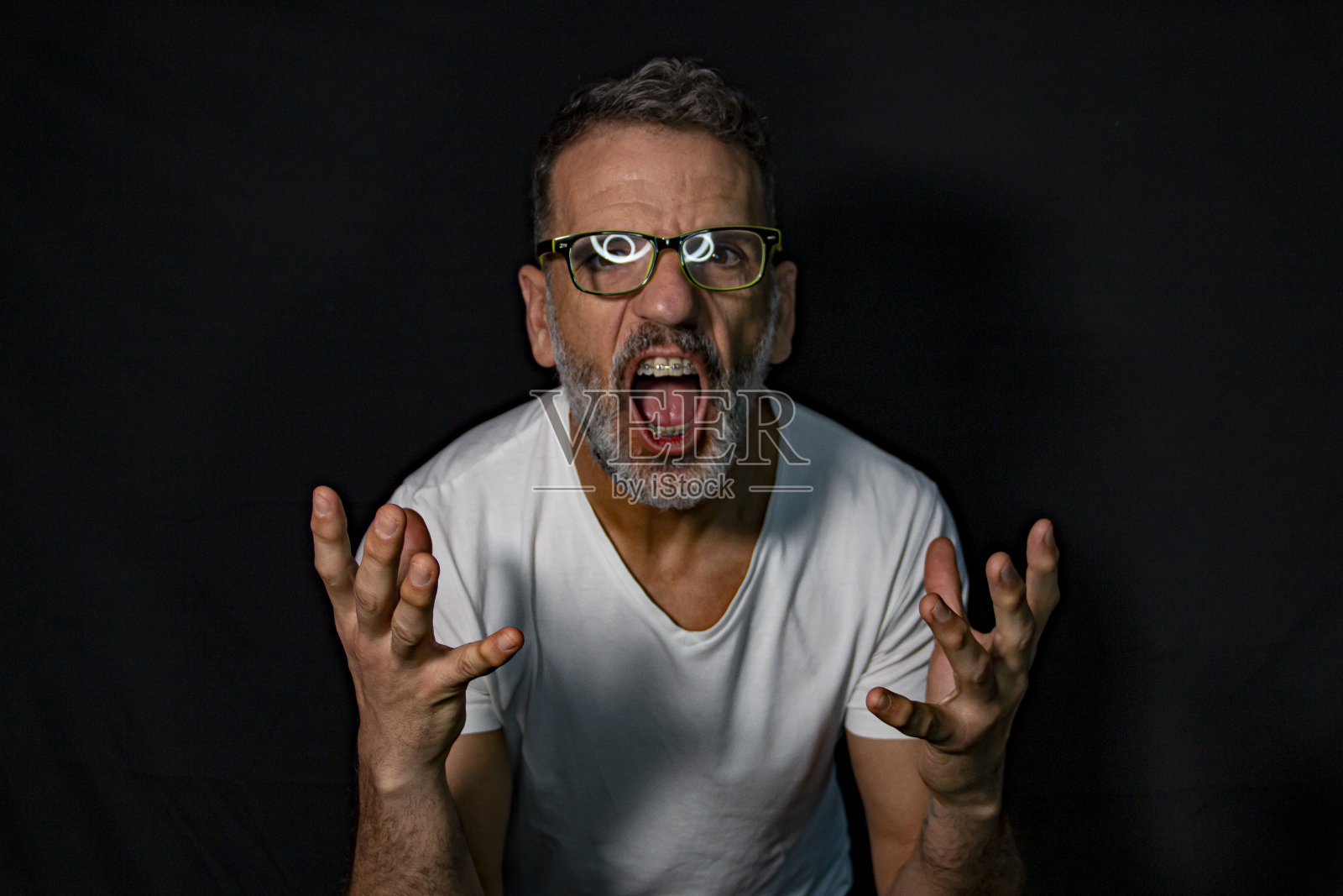一个成熟男人的肖像，留着灰胡子，戴着牙套，用手打手势，穿着黑色背景上的白色t恤。照片摄影图片