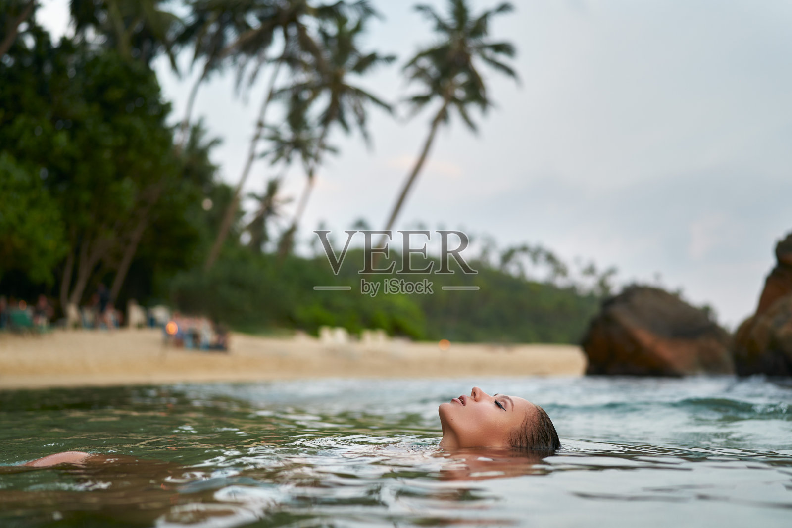 一名女子在热带水域放松，背景是棕榈树。宁静的女性享受防水化妆，而漂浮在海洋。宁静的旅行瞬间，宁静沙滩上时尚的沐浴场景。照片摄影图片