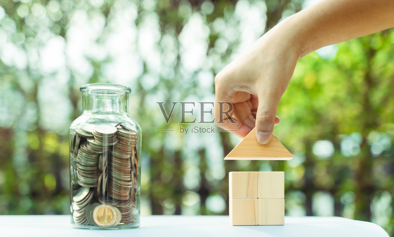 省钱的房子概念，必要的成本的房子，硬币在瓶子和木块房子模型。金钱和财务的房子概念。公园里的散景背景，晨光。照片摄影图片