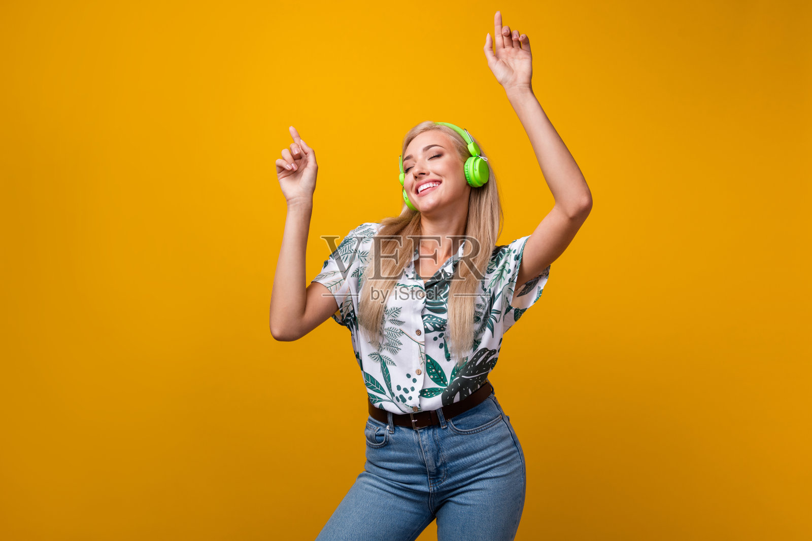 照片中无忧无虑的漂亮女孩，留着长发，穿着衬衫，戴着耳机，在黄色的背景上跳舞，双手举起照片摄影图片