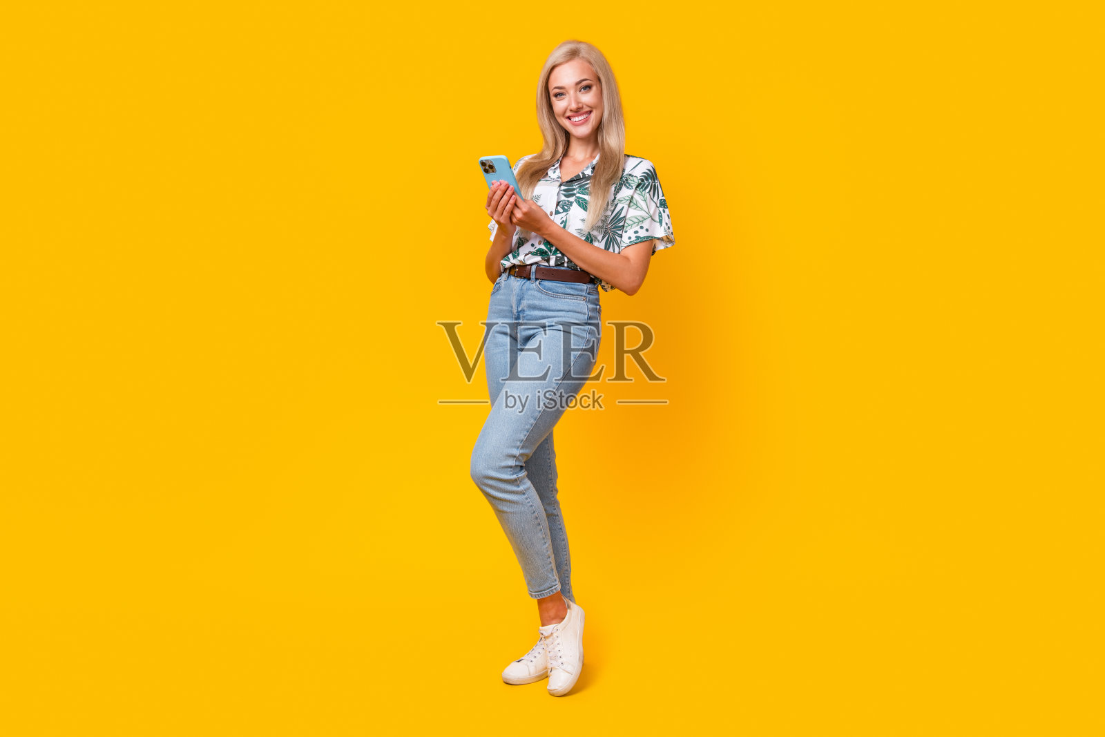 智能漂亮女孩的全尺寸照片，穿着印花衬衫时尚牛仔裤，拿着智能手机孤立在黄色背景上照片摄影图片