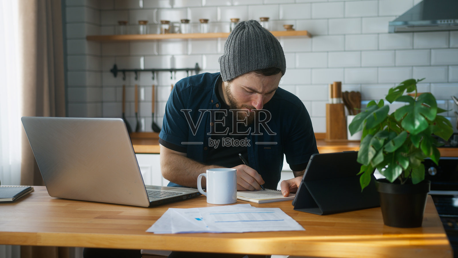 一个戴着无边帽的商人在家厨房里工作用笔记本电脑做笔记，做研究。忙碌的自由职业者在现代科技笔记本设备上工作照片摄影图片