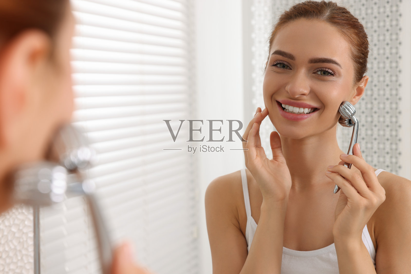 年轻女子在浴室镜子旁用金属滚轮按摩脸部照片摄影图片