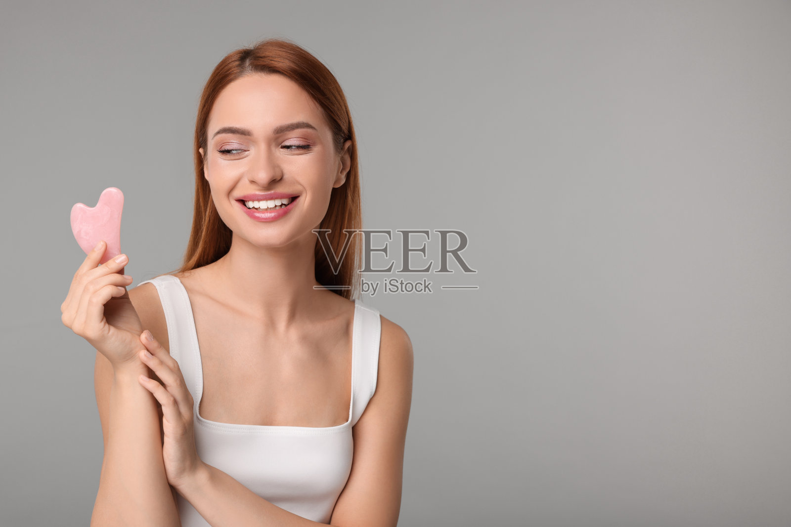 年轻女子用玫瑰石英刨沙工具在灰色背景上，留出文字空间。面部按摩照片摄影图片