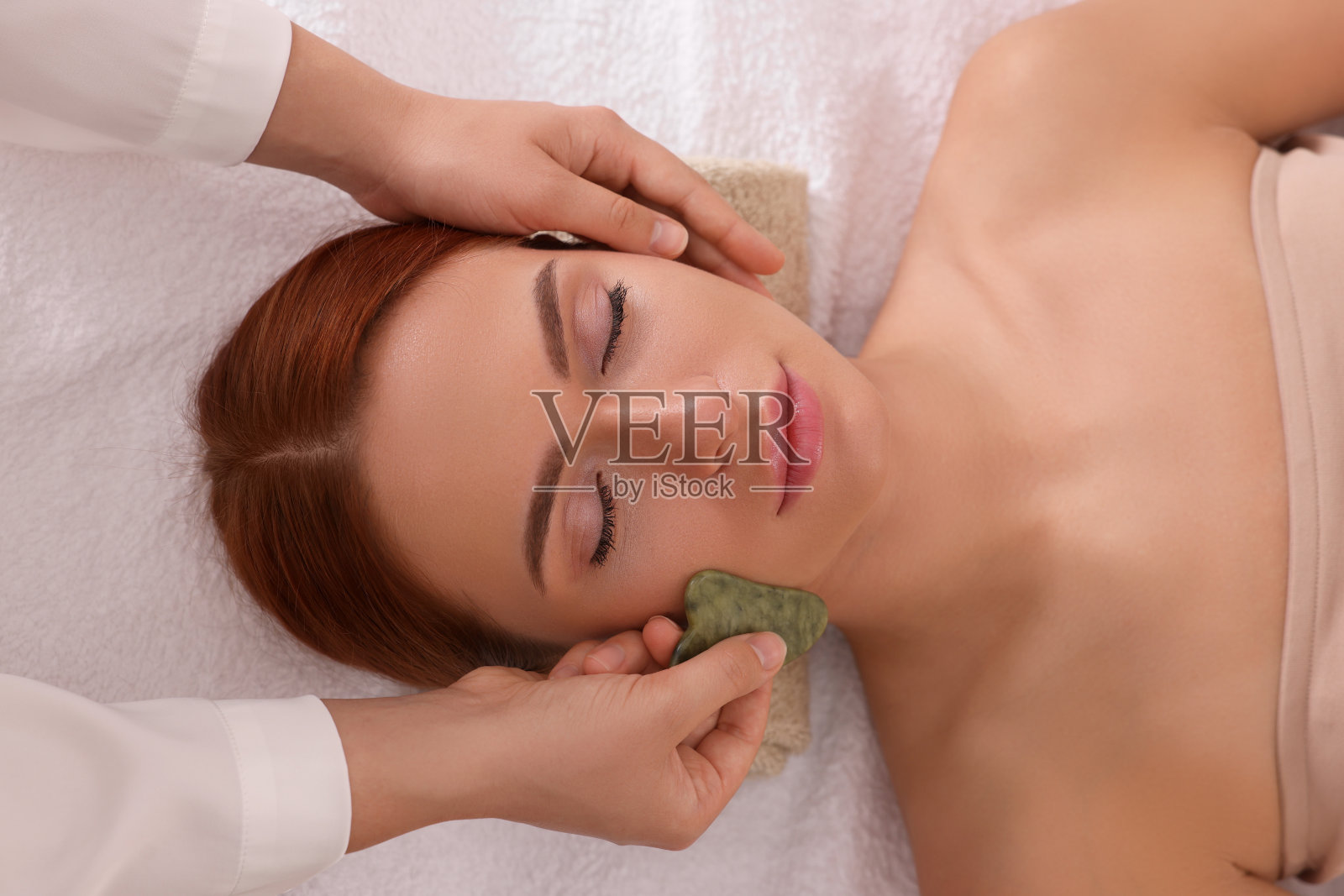 上图，年轻女子在美容院用玉刨沙工具接受面部按摩照片摄影图片