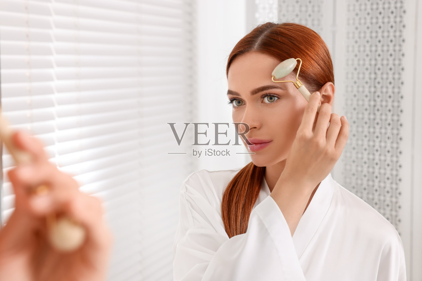 Young woman massaging her face with jade roller near mirror in bathroom照片摄影图片