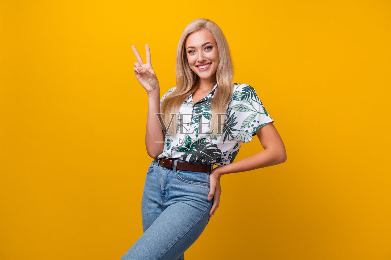 正面女孩的肖像，长发型，穿着印花衬衫，在黄色背景上显示v字手在臀部照片摄影图片